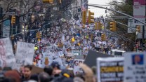 An endless crowd with many holding signs fills a city street, with street lights and lamps overhead.
