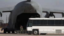 A white bus sits by the open rear hangar of a large gray military aircraft.