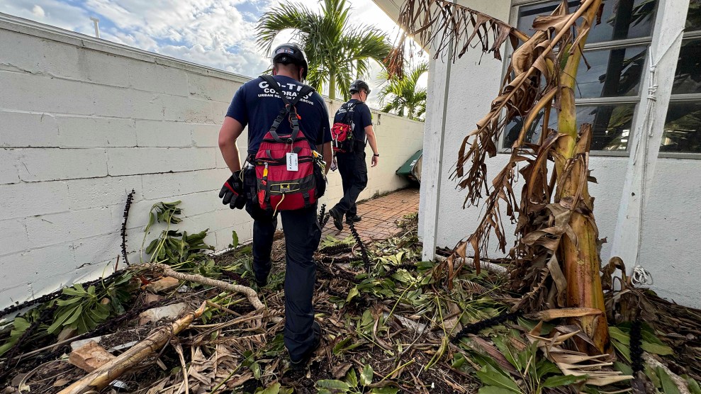 Two people wearing white hard hats and red backpacks walk through a yard full of debris.