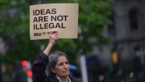 A person with long grey hair holding a sign that says "IDEAS ARE NOT ILLEGAL"