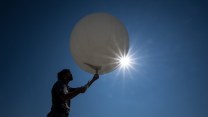 A silhouette figure holds up a white balloon in front of a blues sky.